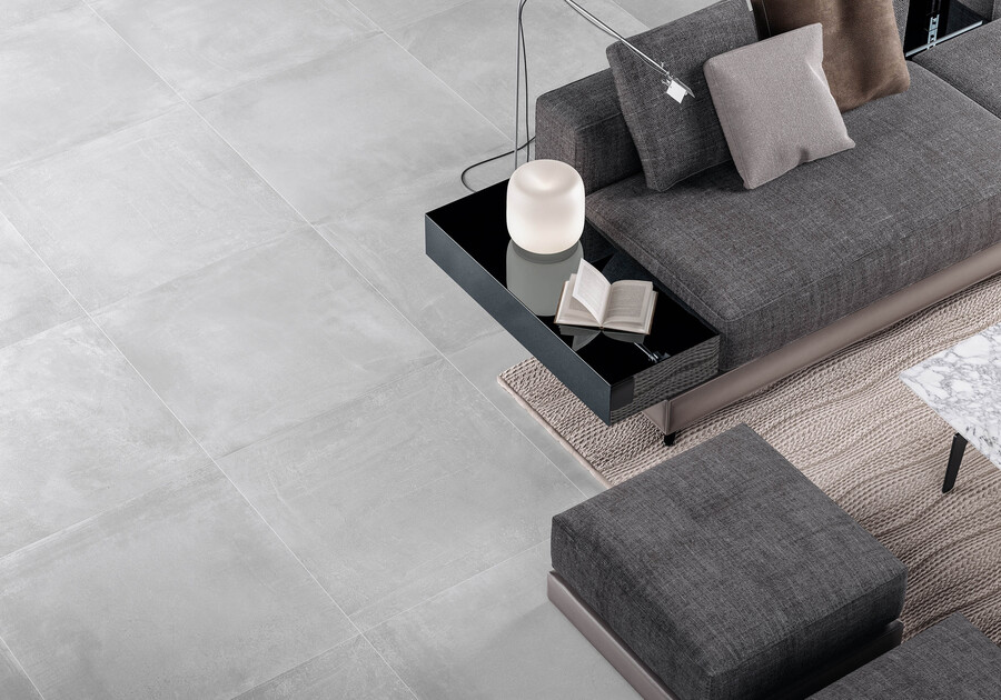 il s'agit d'un séjour avec du carrelage 60x60 cm gris au sol, un canapé gris anthracite, tapis beige et sur la table basse une lampe et un livre ouvert